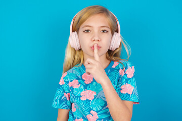 Caucasian kid girl wearing hawaiian T-shirt against blue wall making hush gesture with finger on her lips wearing  wireless headphones. Be quiet.