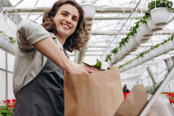 Portrait of male florist selling flowerpot and putting it in eco bag in garden centre. Flower shop.