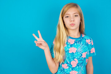 Fototapeta premium Caucasian kid girl wearing hawaiian T-shirt against blue wall makes peace gesture keeps lips folded shows v sign. Body language concept