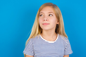 Caucasian kid girl wearing striped shirt ​against blue wall looking aside into empty space thoughtful