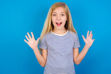 Optimistic Caucasian kid girl wearing striped shirt ​against blue wall raises palms from joy, happy to receive awesome present from someone, shouts loudly, Excited model screaming.