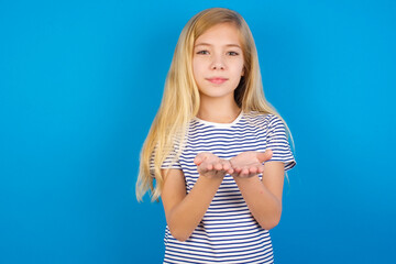 Caucasian kid girl wearing striped shirt ​against blue wall sending blow kiss with pout lips and holding palms to send air kiss.