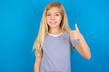 Caucasian kid girl wearing striped shirt ​against blue wall doing happy thumbs up gesture with hand. Approving expression looking at the camera showing success.