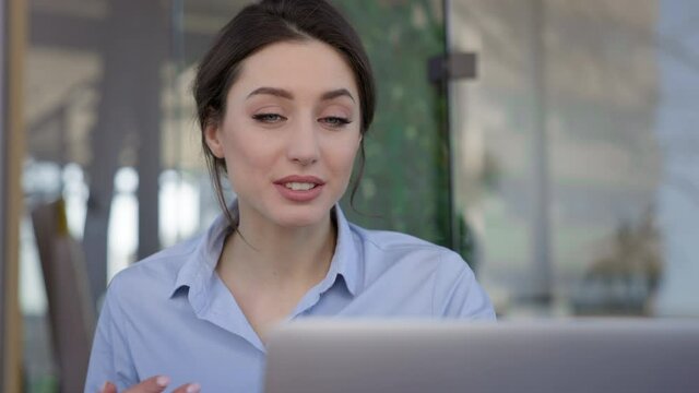 Portrait Of Young Brunette In The Office. She Is Actively Telling Something While Sitting In Front Of Laptop Monitor. The Girl Is Smiling.