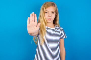 Caucasian kid girl wearing striped shirt ​against blue wall  doing stop sing with palm of the hand. Warning expression with negative and serious gesture on the face.