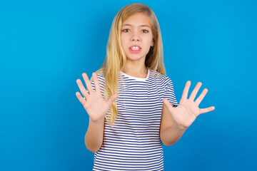 Caucasian kid girl wearing striped shirt ​against blue wall   afraid and terrified with fear...