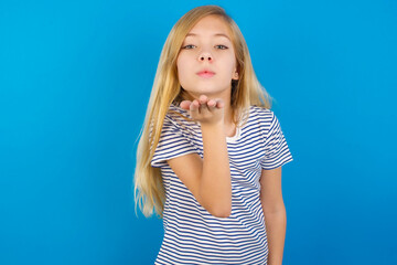 Caucasian kid girl wearing striped shirt ​against blue wall  looking at the camera blowing a kiss with hand on air being lovely and sexy. Love expression.