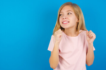 Caucasian kid girl wearing pink shirt against blue wall clenches fists and awaits for something nice happened looks away bites lips and waits announcement of results