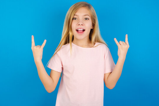 Caucasian kid girl wearing pink shirt against blue wall makes rock n roll sign looks self confident and cheerful enjoys cool music at party. Body language concept.