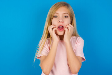 Speechless Caucasian kid girl wearing pink shirt against blue wall keeps hands near opened mouth...