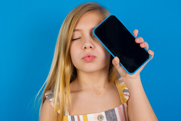Adorable Caucasian kid girl wearing dress against blue wall holding modern device covering eye with lips pouted
