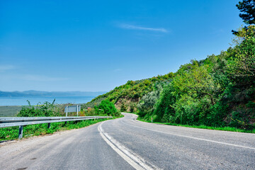 an asphalt road to Bursa to Nicaea (iznik) during sunny day and road signs and protection fences next to the road with small hills that is covered by many green plants and grass with lake of Nicaea.