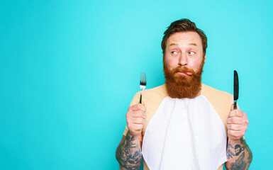 Man with tattoos is ready to eat with cutlery in hand. cyan background