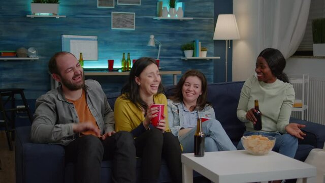 Happy multiethnic friends smiling together while sitting in frot of television looking at funny comedy movie. Group of mixed race people relaxing on couch late at night in living room