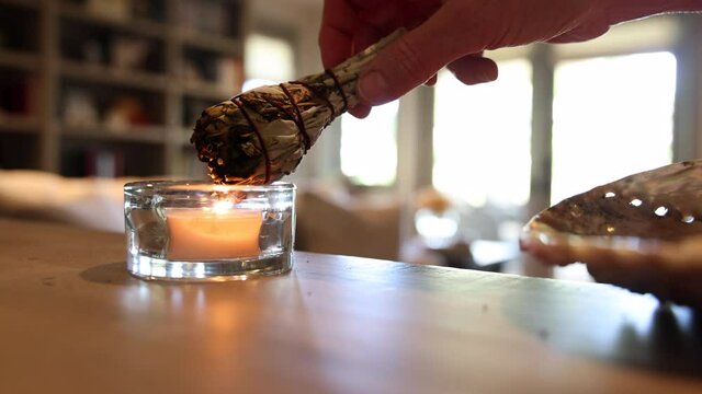 Hand of woman picking up wrapped sage bundle then bringing it to a candle to light it and waving smoke through home with a feather