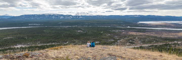 Fototapeta na wymiar Two people, couple hikers sitting on a panoramic view above Yukon River outside of Whitehorse in north of Canada. Boreal forest, snow capped mountains on a cloudy afternoon in distance. 