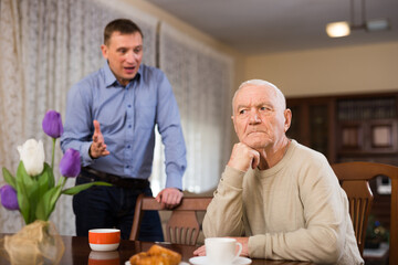 Offended elderly man sitting at home table with disgruntled adult son standing behind and reprimanding him