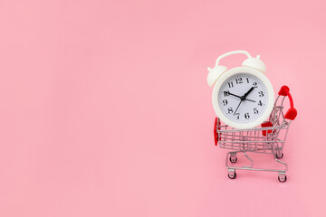 White alarm clock lies in the shopping cart on pink background. Big sale time concept. Copy space.