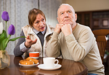 Fototapeta na wymiar Offended elderly man sitting at home table on background with angry wife reprimanding him