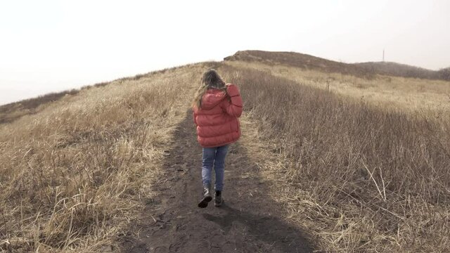 Walk in the field. Back view. A girl in a red jacket walks along a narrow path next to a dry field. A child walks in a field with tall dry grass.