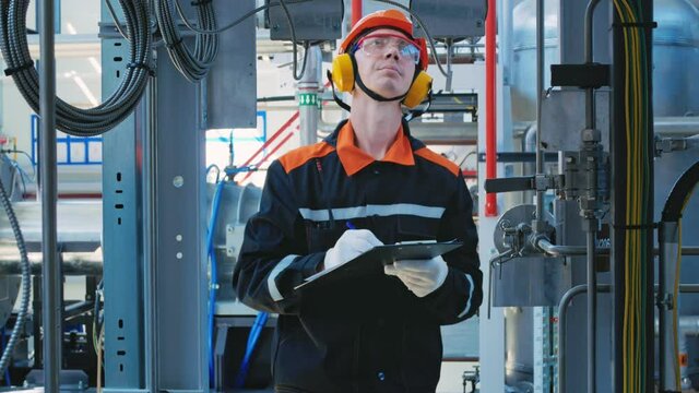 An engineer technician in a helmet, goggles and headphones, writes down data on the operation of technological equipment with a pen on a sheet of paper. Work in industrial. Manufacturing industry.