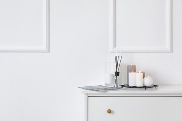 Burning candles, reed diffuser and magazine on chest of drawers near light wall