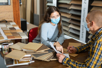 Experienced salesperson in protective face mask working with client offering laminate flooring in shop of building materials