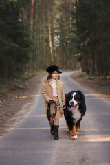 little girl in a coat and shorts and a cap walks in the forest with a bernese mountain dog