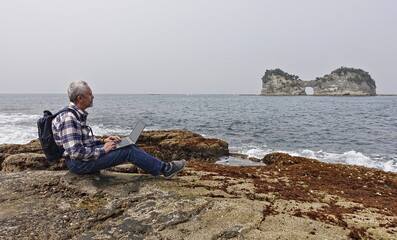 gray-haired man in jeans and a plaid shirt works with a laptop on the ocean shore