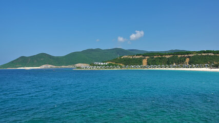 Summer seascape. There are ripples in the turquoise water. A row of villas can be seen on the sandy beach. Green mountains against the blue sky. Vietnam.