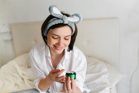  woman holding a green jar of cream in her hands, Brunette girl in  real bedroom interior applies cream on her face  facial skin care, time for herself and cosmetology, without retouching