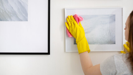 Home cleaning. Janitor woman. Housework routine. Unrecognizable woman in yellow protective gloves clearing picture art from dust microfiber cloth.
