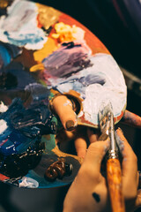 Art school. Creative education. Supplies tools. Blue white orange acrylic paint on palette brush in professional female artist hands on dark background.