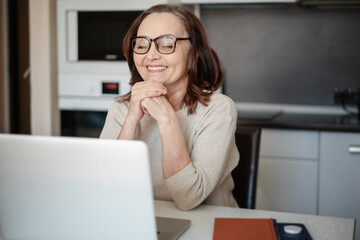 Serious senior woman in glasses working at home with a laptop while sitting in the kitchen.