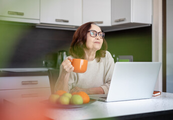 A senior woman in glasses working at home with a laptop and drinking coffee from a big mug sitting in the kitchen.