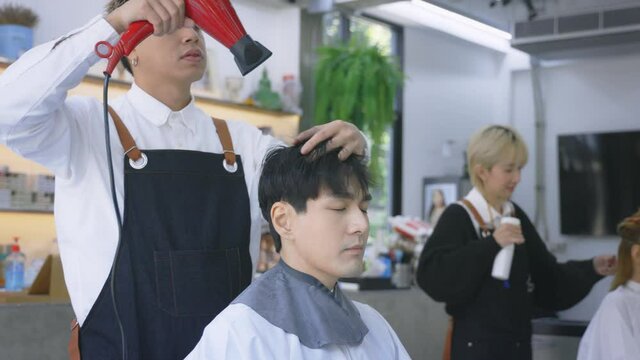 Young male Asian hairdresser wearing apron drying handsome young man's hair in a modern barber shop while concentrating on work