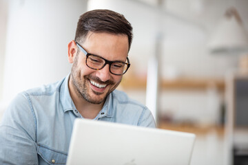 Young man working at home in the evening. Young businessman working at home using lap top. Young attractive smiling guy is browsing at his laptop, sitting at home