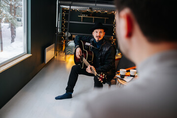Fototapeta na wymiar Bearded middle aged manin a hat sitting with guitar in hands at home. He is wearing casual clothes and leather jacket atop. looking at a man in foreground