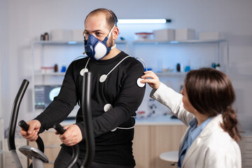 Man athlete with mask running on cross trainer after medical researcher attached electrodes to her...