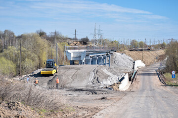 Construction equipment forms an artificial embankment and compresses the ground against the background of the construction of a new bridge.