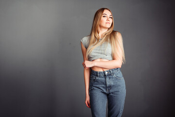 Long-haired blonde in jeans against the background of a gray wall hugs herself and smiles sincerely