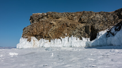 Fototapeta na wymiar A picturesque granite rock, devoid of vegetation, against the backdrop of a blue sky. There are cracks on the stones, the base is covered with ice splashes, icicles. Around is a frozen lake. Baikal