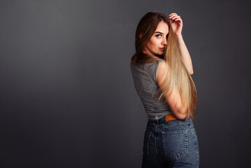 European girl in a gray top and jeans with blond long hair, stands sideways and holds her hand behind her head, looks directly at the wall