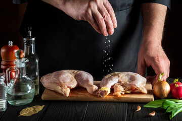 Сook or chef adds salt to the raw chicken legs. Preparation for grilling with chicken leg in the kitchen of a restaurant or cafe on a table with vegetables and spices