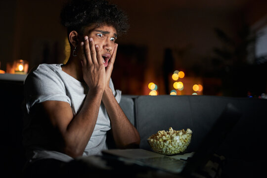 Young Guy Looking Terrified While Watching Horror Movie Alone Using Laptop And Eating Popcorn, Sitting On A Comfortable Sofa In Dark Room At Home