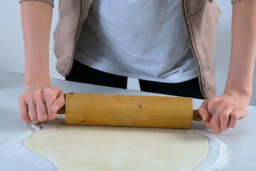 Woman is rolling dough for dumpings using rolling pin on table in kitchen, hands closeup. Process of cooking dumplings at home. Minced meat, dough and rolling pin on the table.