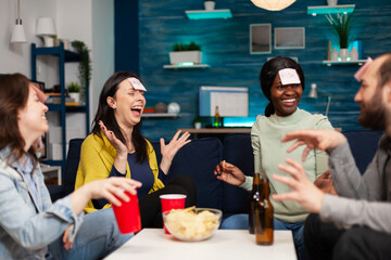 Mixed race friends playing board game using sticky notes spending time together late at night relaxing on couch in living room. Group of multi-ethnic people drinking beer, eating chips enjoying home