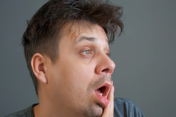 Fototapeta premium Tired boring shaggy young man on grey background. He is keeping his head by hands. Depression, apathy, and mental illness in the modern world. Middle age crisis.