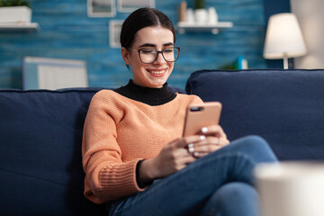 Relaxed student sitting on couch in living room while writing message holding modern smarphone in hands. Teenager woman chatting on social network looking at phone screen - obrazy, fototapety, plakaty