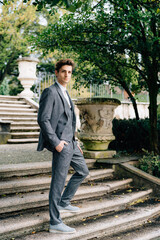 Fototapeta na wymiar Serious groom stands with his hands in his pockets on the stairs in the park near the old villa. Lake Como